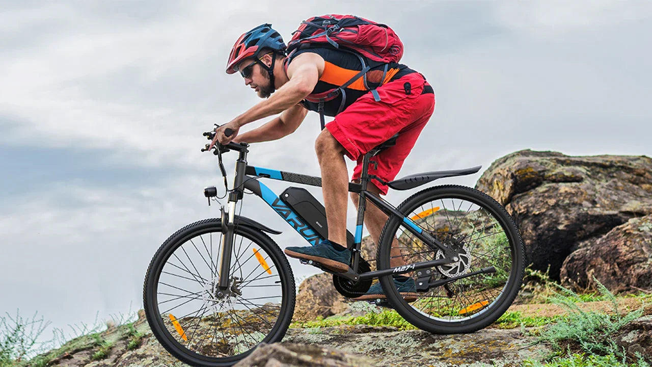 Person riding a VARUN M27-1 Electric mountain bike on rocky terrain with a backpack and helmet.